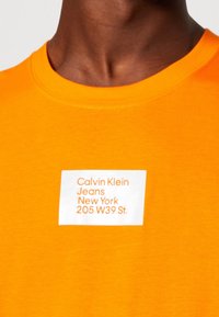 Persona que lleva una camiseta naranja brillante con una etiqueta rectangular blanca que muestra "Calvin Klein Jeans New York 205 W39 St." en texto naranja.