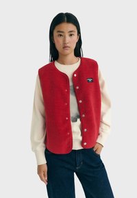 Rode fleece vest met drukknoopjes, ronde halslijn en logopatch, gedragen over een lichtgekleurde sweatshirt en donkere spijkerbroek.