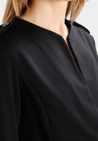 Svart klänning med hög halsringning, mjukt strukturerad silhuett och knappdetaljer på axlarna. Slät tygstruktur.