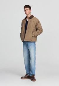 Giacca di cotone marrone con colletto marrone scuro, abbinata a jeans blu e scarpe marroni. Il modello sta in piedi con le mani in tasca contro uno sfondo neutro.