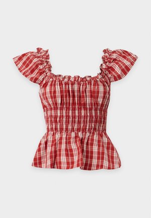 Vrouw draagt een rode geruite top met ruches en zwarte wijde broek, houdt een grote zwarte leren handtas vast, staand tegen een effen achtergrond.