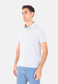 Polo shirt grigio chiaro a maniche corte con colletto e pattina con tre bottoni; realizzato in un tessuto morbido e strutturato. Indossato con jeans blu.