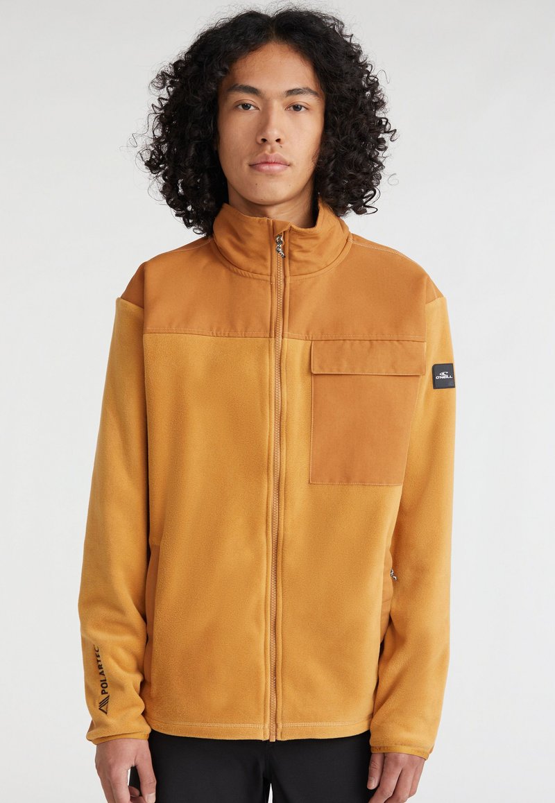 Fleece jakna v gorčični rumeni barvi z visokim ovratnikom, sprednjim zadrgo, žepom na prsih in logotipom na rokavu. Mehka tekstura in preprost design.