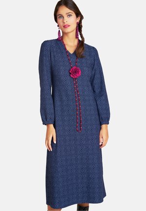 Langes, marineblaues Kleid mit strukturiertem Muster, Ballonärmeln und V-Ausschnitt. Akzentuiert mit einer langen Perlenkette und einem Blumendetail.