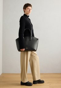 Black leather tote bag with a structured design, dual handles, and a triangular base. Features subtle gold branding and a smooth texture.