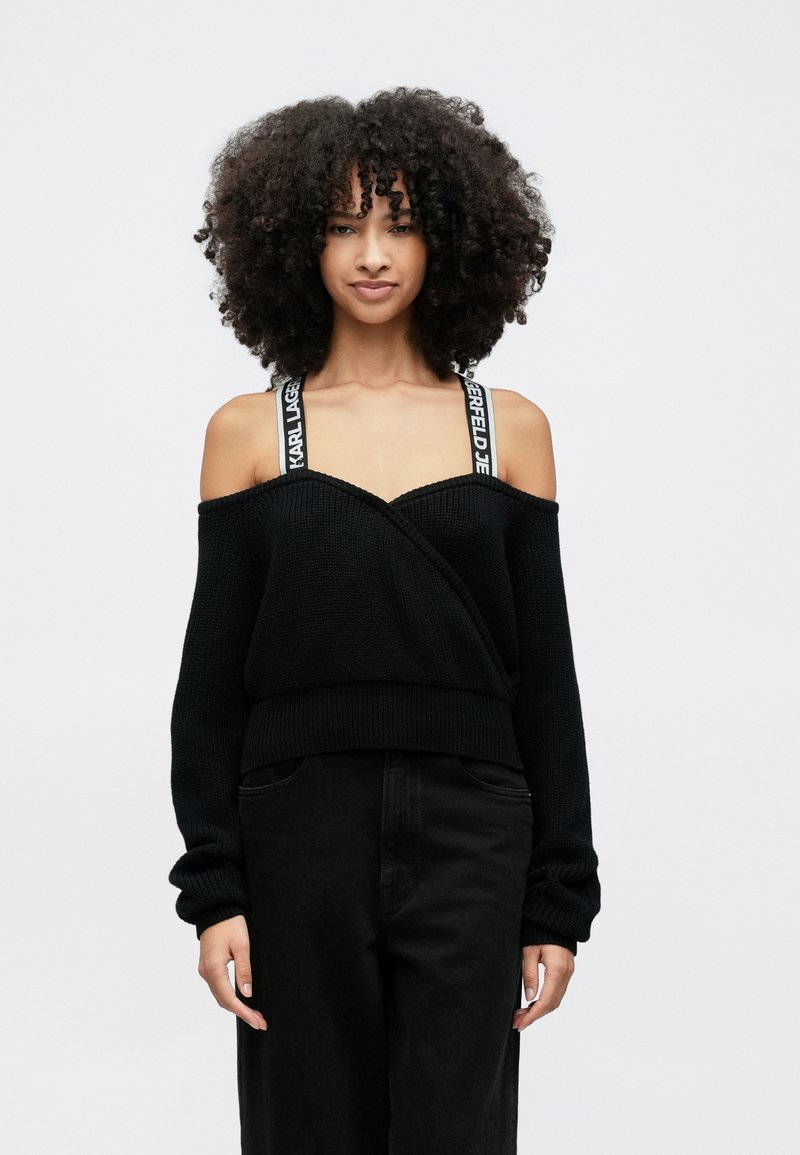 Kvinna med lockigt hår som bär en svart stickad tröja med bara axlar, synliga märkta band och svarta byxor, stående mot en enfärgad vit bakgrund.