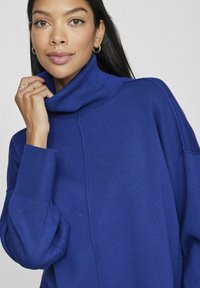 Zils augstais kakls džemperis ar ribotu tekstūru, brīvs piegriezums un nolaistas plecu šuves. Iespējas garas piedurknes un smalka šuvju detalizācija.