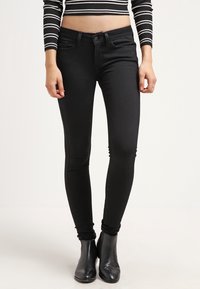 Vaqueros skinny negros con una textura suave, diseño de cinco bolsillos y bucles para cinturón, combinados con botas negras hasta el tobillo y una blusa acortada de rayas.