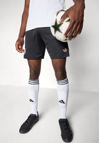 Svarte treningsshorts med en gulllogo, kombinert med hvite sokker med svarte striper og en fotball med grønne og svarte aksenter.