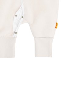 Babysoverall aus weichem beigem Stoff mit weißen Druckknöpfen und Bündchen. Beinhaltet ein kleines gelbes Etikett an der Seite für Branding und erleichtertes Ankleiden.