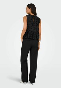 Femme portant un haut noir sans manches texturé avec un motif floral et un pantalon large noir, debout, les mains dans les poches, de dos.