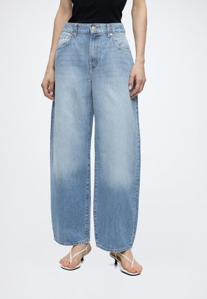 Person iført lyseblå jeans med brede ben, sort top og hvide sandaler, stående mod en ensfarvet hvid baggrund.