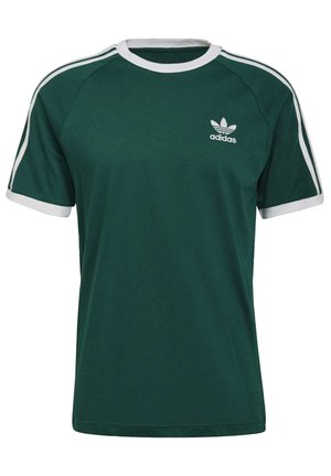 Mand iført mørkegrøn Adidas t-shirt med hvide striber på ærmerne og Adidas-logo på brystet, kombineret med sorte bukser.