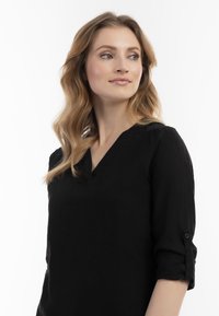 Sort v-hals bluse lavet af tekstureret stof med rullede ærmer sikret af knapdetalje. Har en enkel, klassisk silhuet.