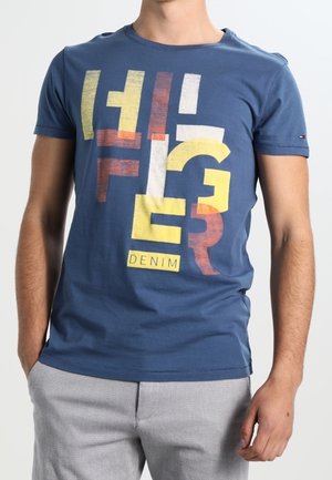 Homme portant un t-shirt bleu à manches courtes avec des lettres abstraites jaunes, rouges, blanches et le mot "DENIM", associé à un pantalon gris clair.