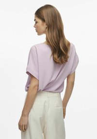 Gaiši violets īsām piedurknēm tops ar brīvu piegriezumu, teksturētu audumu un noapaļotu dekoltē, kombinēts ar bežām augstiem stulpiem.