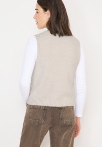 Hellgraue gestrickte Weste mit strukturiertem Finish, geripptem Saum und ärmellosem Design, kombiniert mit braunen Denim-Hosen.