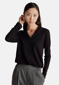 Czarny long-sleeve top z dekoltem w kształcie V, wykonany z gładkiego materiału. Modelka ma na sobie szare, plisowane spodnie, trzymając ręce na szyi.