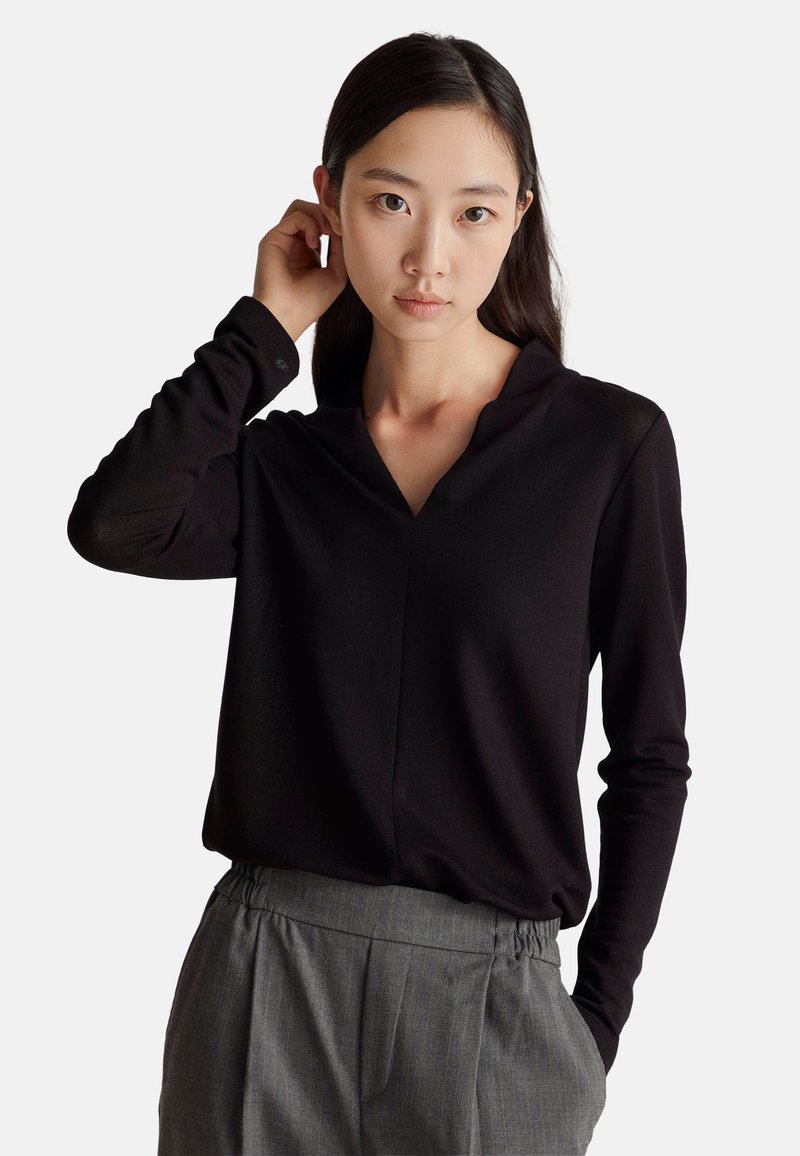 Czarny long-sleeve top z dekoltem w kształcie V, wykonany z gładkiego materiału. Modelka ma na sobie szare, plisowane spodnie, trzymając ręce na szyi.