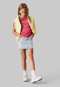 Camiseta rosa de Calvin Klein, falda de mezclilla azul clara, suéter amarillo sobre los hombros, zapatillas blancas y calcetines blancos de tobillo.