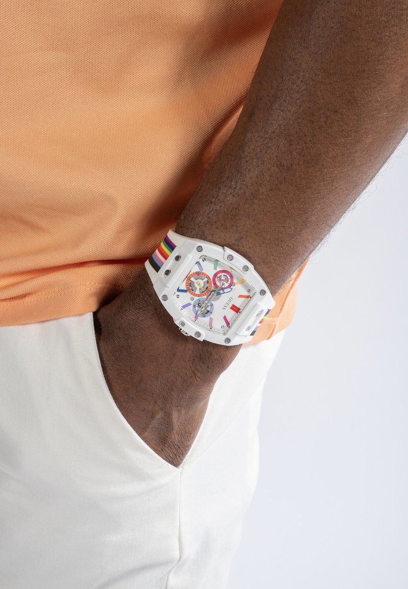 Bras à la peau foncée avec la main dans la poche d'un pantalon blanc, portant une montre blanche avec des détails aux couleurs de l'arc-en-ciel et une chemise orange.