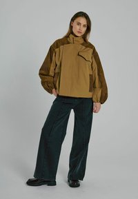 Braune übergroße Jacke mit hohem Kragen und einer Klappentasche, mit strukturierten Ärmeln und kombiniert mit weiten schwarzen Jeans und schwarzen Schuhen.