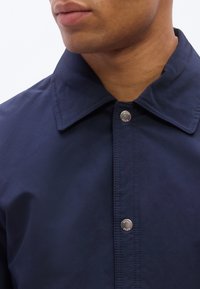 Κοντινό πλάνο ατόμου που φορά μπλε μπλούζα με γιακά και εμφανή ασημένια κουμπιά κατά μήκος της λωρίδας  κουμπιών.