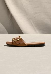 Sandale mule en cuir marron avec une boucle rectangulaire dorée et une semelle plate noire sur une surface neutre.