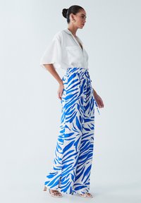 Chemise col blanc, assortie à un pantalon à taille haute et à jambes larges avec un motif de feuilles bleu et blanc. Le modèle porte des sandales blanches.
