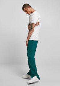 Balta īsā piedurkņu T-kreklā, teal krāsas sporta biksēs ar rāvējslēdzēju akcentiem un baltos sporta apavos. Redzams tetovējums uz apakšdelma. Neitrāls pelēks fons.