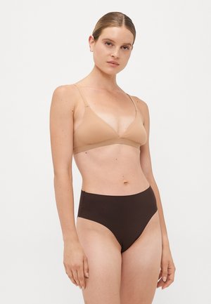 Woman with blonde hair in a nude triangle bra and high-waisted dark brown underwear standing against a plain, light background.