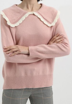 Mujer con un suéter rosa con un gran cuello blanco con borde en forma de concha y brazos cruzados, combinado con pantalones a cuadros en blanco y negro.