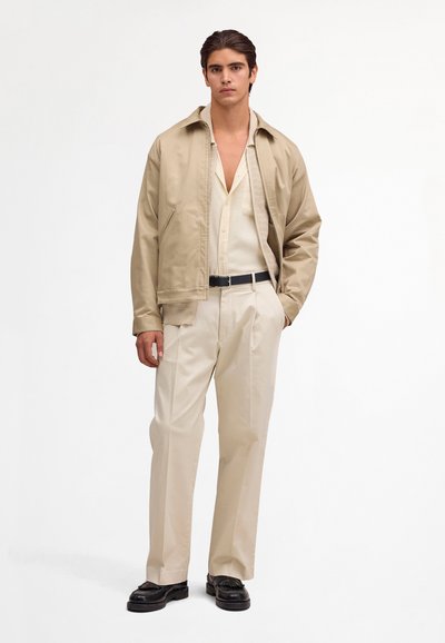 Jeune homme debout portant une veste beige, une chemise crème et un pantalon, une ceinture noire et des mocassins noirs, sur un fond blanc uni.