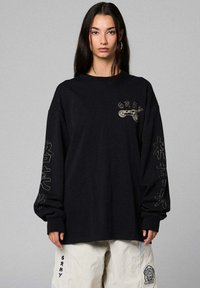 Vrouw met lang donker haar, gekleed in een zwart oversized shirt met lange mouwen met geborduurde tekst en afbeeldingen, en witte broek met vlekken.