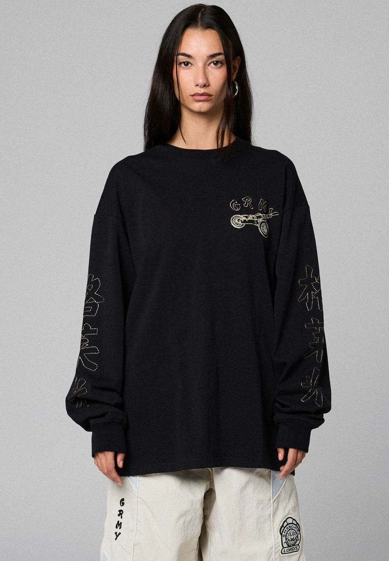 Vrouw met lang donker haar, gekleed in een zwart oversized shirt met lange mouwen met geborduurde tekst en afbeeldingen, en witte broek met vlekken.