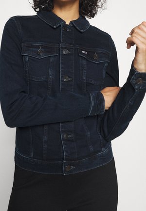 Osoba ubrana w ciemnoniebieską kurtkę dżinsową z zapinanymi kieszeniami i guzikami z przodu, ze skrzyżowanymi rękami na piersi.