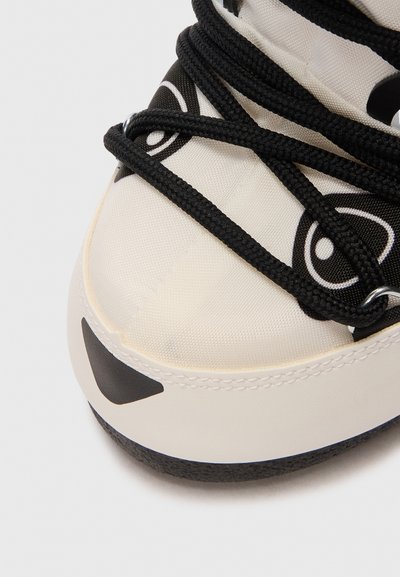 Close-up van de neus van een witte sneaker met een zwart oogvormig ontwerp en zwarte veters die over de stoffen bovenkant kruisen.