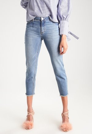 Frau trägt hellblaue, gekürzte Jeans mit ausgefransten Saum, eine blau-weiß gestreifte Bluse mit gebundenen Manschetten und nudefarbene High Heels mit rosa Federdetails.