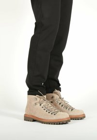 Persona que lleva botas de senderismo de ante beige con ojales de metal y suelas de goma gruesa, combinadas con pantalones negros sobre un fondo blanco.