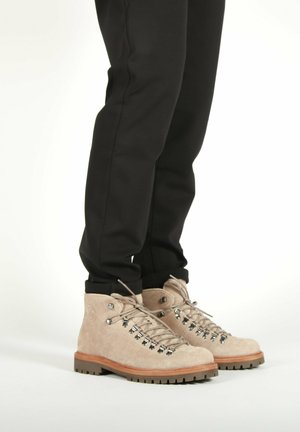 Persona que lleva botas de senderismo de ante beige con ojales de metal y suelas de goma gruesa, combinadas con pantalones negros sobre un fondo blanco.