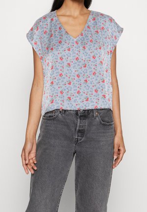 Kvinde iført lyseblå V-hals bluse med rødt blomsterprint og sorte højtaljede jeans, stående foran en helt hvid baggrund.