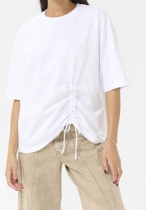 Osoba ubrana w białą, oversize'ową koszulkę z marszczeniem z przodu oraz beżowe spodnie o szerokich nogawkach, z widoczną złotą bransoletką i pierścionkami na palcach.