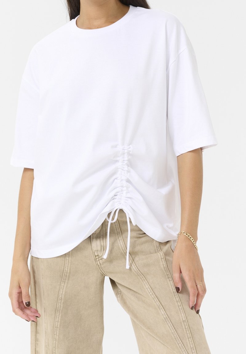 Personne portant un t-shirt blanc oversize avec fronces à l'avant et un pantalon ample beige, bracelet en or et bagues visibles aux doigts.