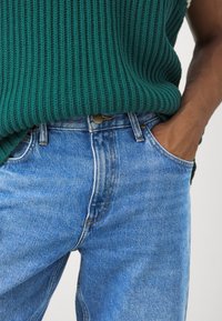 Pulover verde cu model ribat pe deasupra unor blugi din denim de un albastru deschis. Blugii au o croială clasică, cusături vizibile și un nasture cu marcaj.