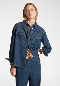 Camicia di jeans in lavaggio scuro con vestibilità rilassata, dettagli in studs lungo il colletto e le tasche, e chiusura con bottoni. Abbinata a un capo intimo a fantasia.