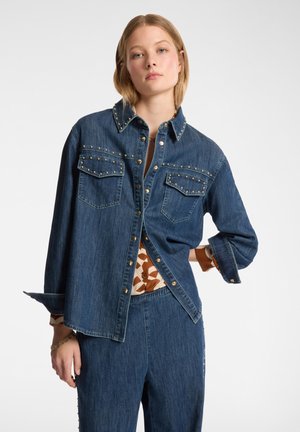 Camisa de mezclilla en lavado oscuro con un ajuste relajado, acentos de tachuelas en el cuello y los bolsillos, y cierre de botones. Combinada con una prenda interior estampada.