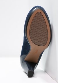 Námořnické modré semišové lodičky s špičatou špičkou a černou koženou panely. Texturovaná gumová podrážka s zakřiveným podpatkem. Viditelné logo značky.