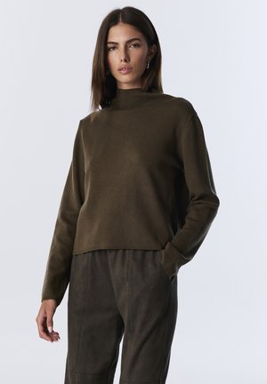 Jeune femme aux cheveux bruns raides portant un pull col montant olive et un pantalon assorti, se tenant avec une main dans sa poche devant un fond blanc.