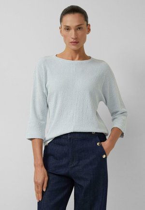 Kobieta z zaczesanymi do tyłu włosami, ubrana w jasnoniebieską teksturowaną bluzkę z rękawem 3/4 oraz ciemne, wysokie spodnie z złotymi guzikami, pozuje z jedną ręką w kieszeni.