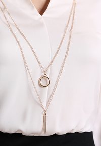 Lagererad halskedja i guldton med en cirkulär berlock och en vertikal stänger, satt på en delikat kedja med en slät finish.